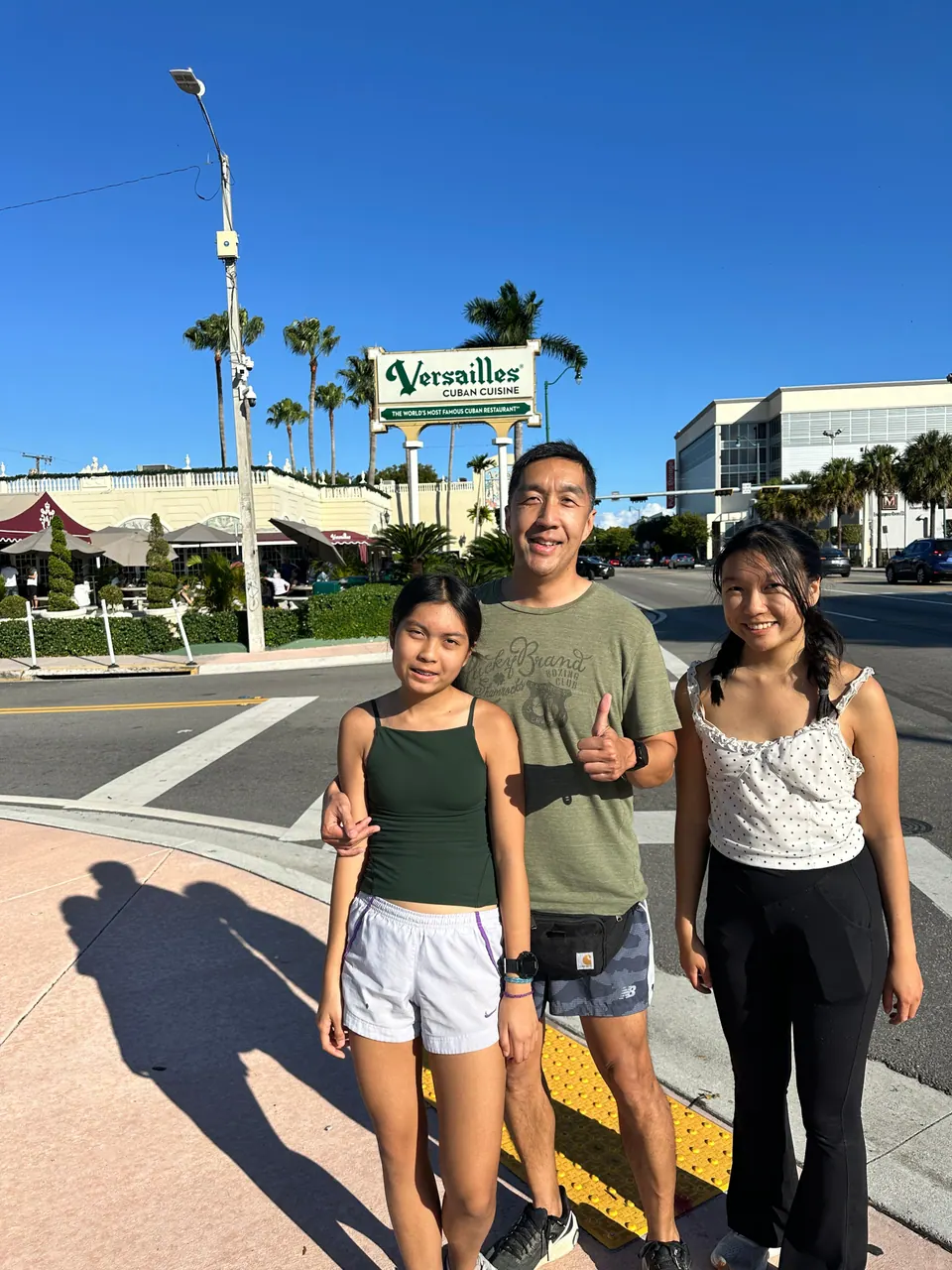 A man in an olive T-shirt gives a thumbs-up between two girls at a crosswalk, in front of the green Versailles Cuban Cuisine road sign and palm trees.