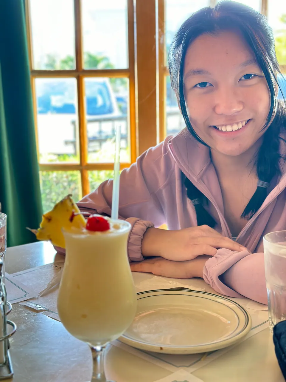 A girl in a pink hoodie smiles across a white-clothed table at a piña colada topped with a pineapple wedge and a cherry, beside a tall window with green curtains.