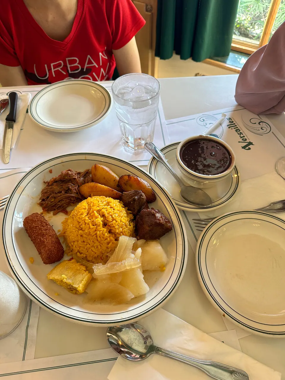 A white plate with crested china holds shredded beef, yellow rice, a croquette, plantain and yuca, with a small cup of black bean soup beside it.