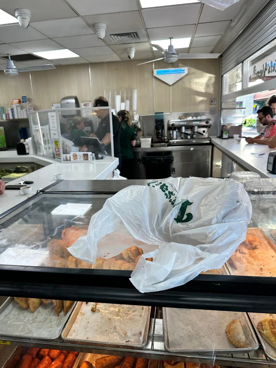 Behind a glass pastry case, with an espresso machine and staff at work, a white takeaway bag sits on top of the case full of golden pastries.
