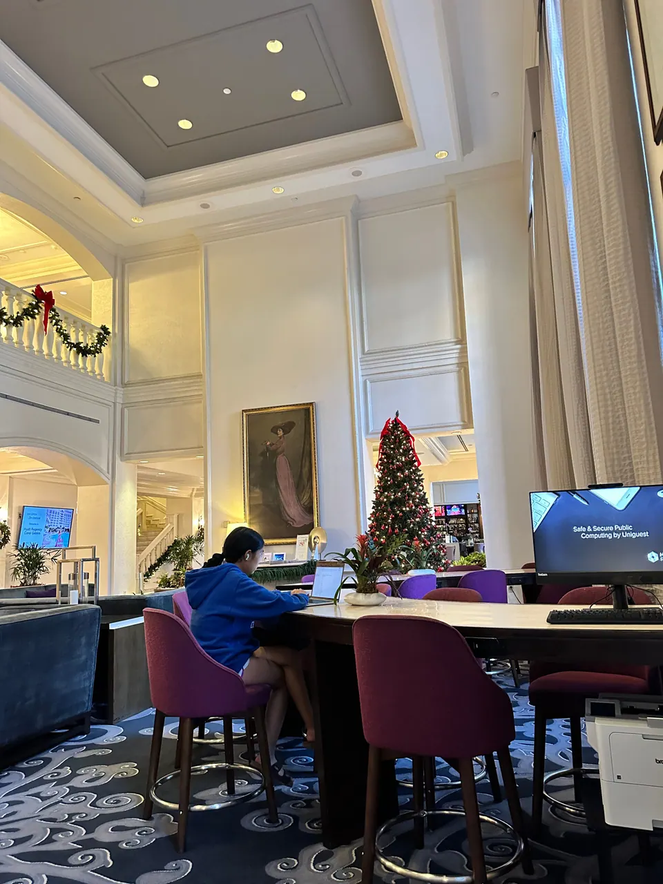 A girl in a blue hoodie sits at a long lobby desk with a laptop, in a tall room with a Christmas tree, a garlanded balcony and a wall screen behind her.