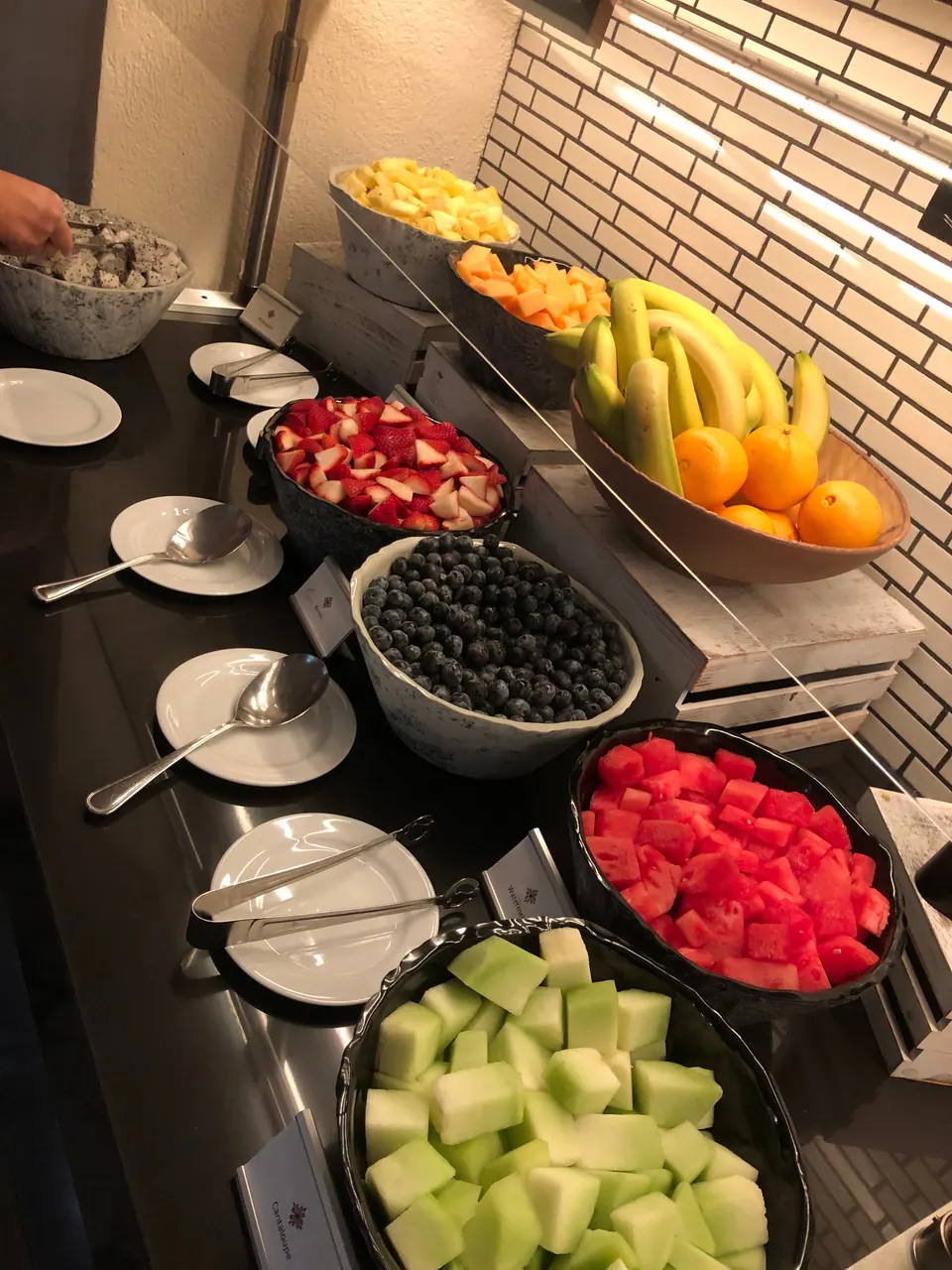 A fruit bar in a hotel restaurant holds bowls of watermelon, honeydew melon, blueberries, strawberries and other fruit, with bananas and oranges on a stand above.