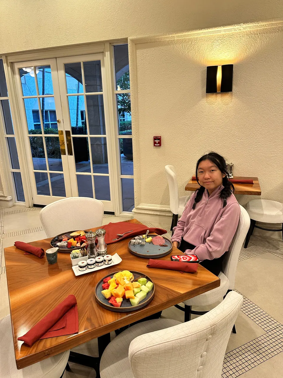 A girl in a lilac hoodie sits at a wooden table with red napkins and plates of fruit and small dishes, beside tall arched glass doors.