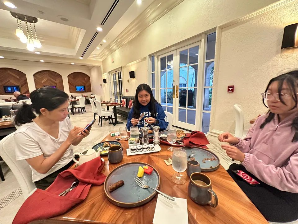 Three people sit at a round wooden table with red napkins, coffee cups and plates in a restaurant with arched doors and windows.