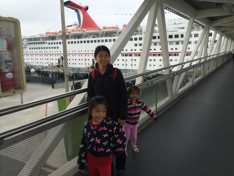A woman in a black fleece stands on a covered walkway of white steel beams with two small girls, one in a butterfly-print jacket and one in a striped top; a large white cruise ship is moored behind them.