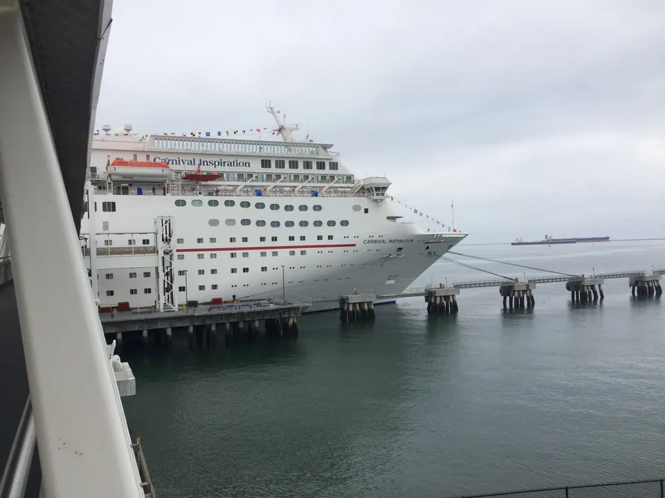 A large white cruise ship with the name Carnival Inspiration lettered on its hull is moored at a long pier, seen past the white steel beams of a gangway under a gray sky.