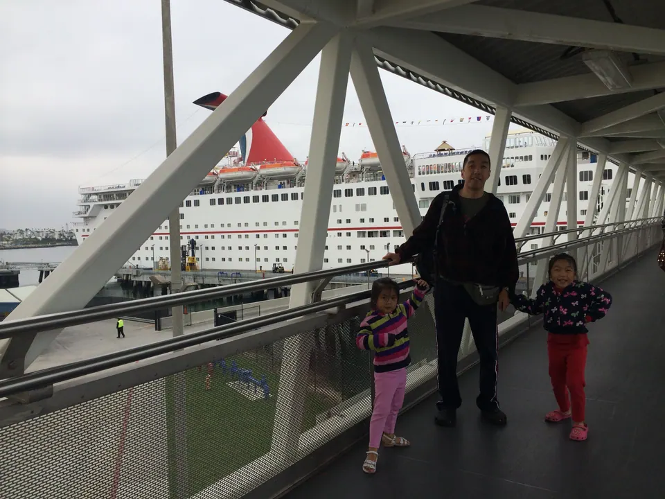 A man holds hands with two small girls on a covered walkway of white steel beams; a large white cruise ship with a red winged funnel is moored behind them.