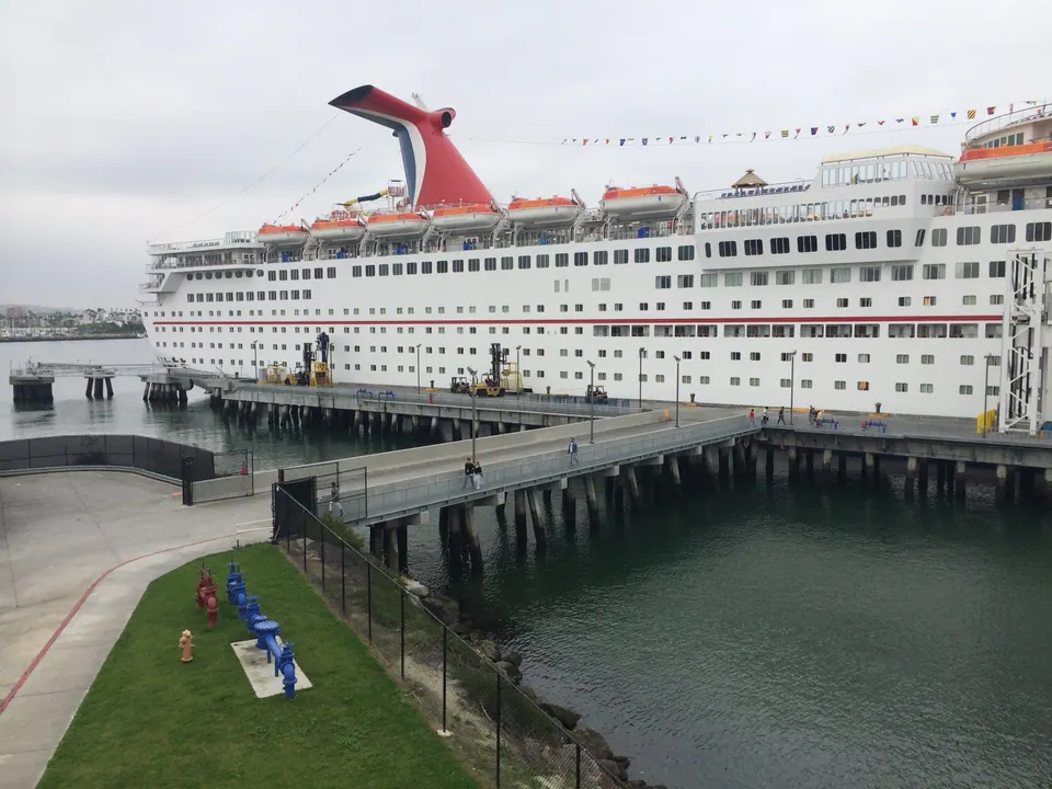 A large white cruise ship with a red, white and blue winged funnel and orange lifeboats is moored along a pier, with a strip of lawn and harbor water in front.