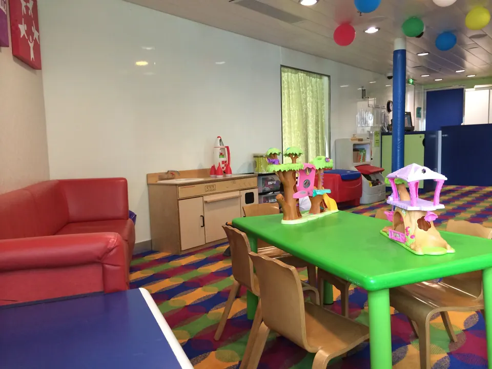 A children's playroom with a bright green table, toy tree houses, a red sofa, a rainbow carpet and balloons hanging from the ceiling.
