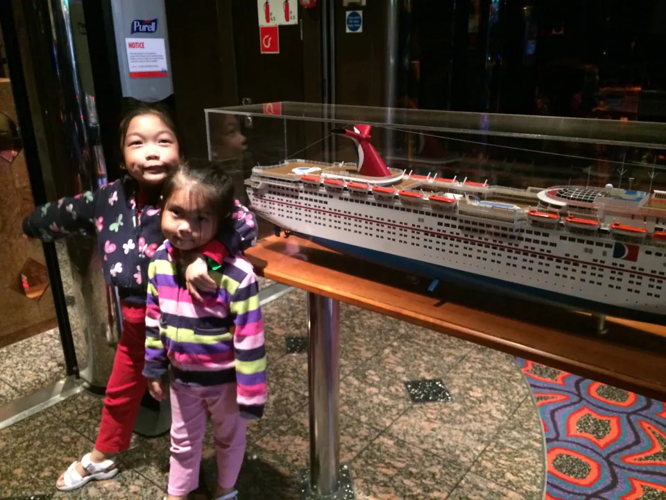 Two small girls, one in a butterfly-print jacket and one in a striped top, stand beside a scale model of a cruise ship in a glass case on a wooden stand, on a patterned carpet.