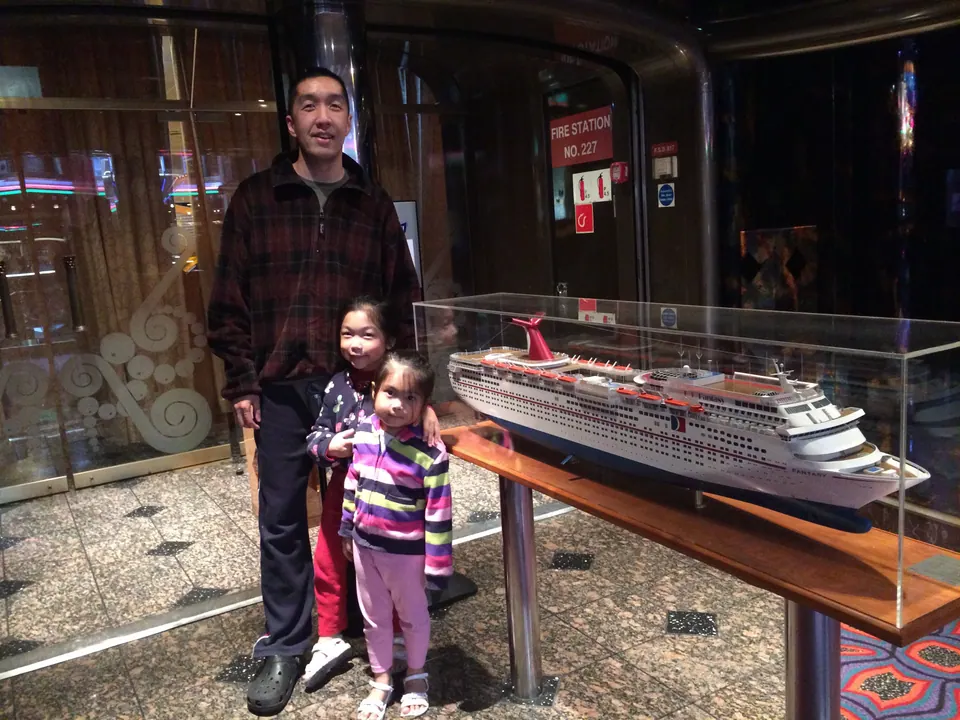 A man in a plaid shirt stands with two small girls beside a glass case holding a model of the ship, in a dark wood-panelled lobby.