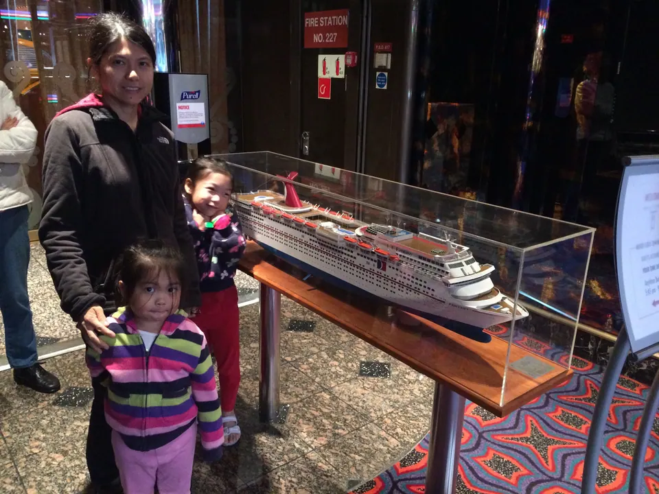 A woman stands with two small girls beside a scale model of a cruise ship in a glass case in a lobby with patterned carpet.