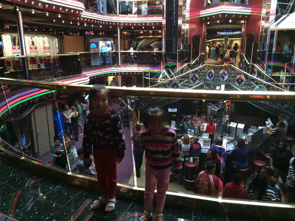Two small girls lean at a glass balcony rail above the ship's atrium, with tiers of shops, neon-lit balconies and crowds of passengers below.