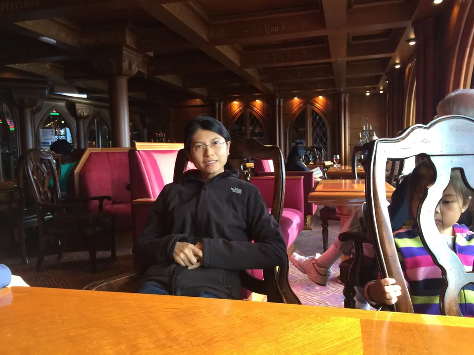 A woman in a black fleece sits at a wooden table in a dark wood-panelled lounge with round windows and pink upholstered chairs.