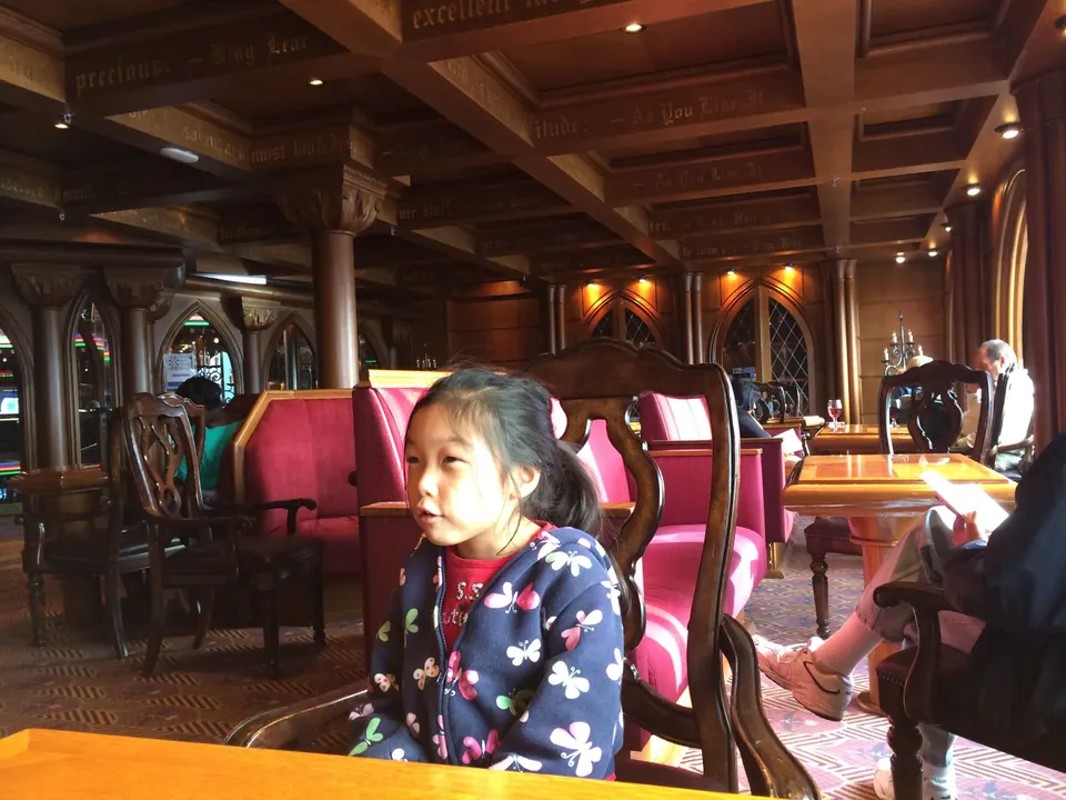 A girl in a navy butterfly-print jacket sits on a pink chair in an almost empty wood-panelled lounge, looking off to one side.