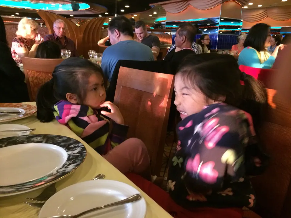 Two small girls lean toward each other laughing at a dinner table set with white plates, in a busy dining room.