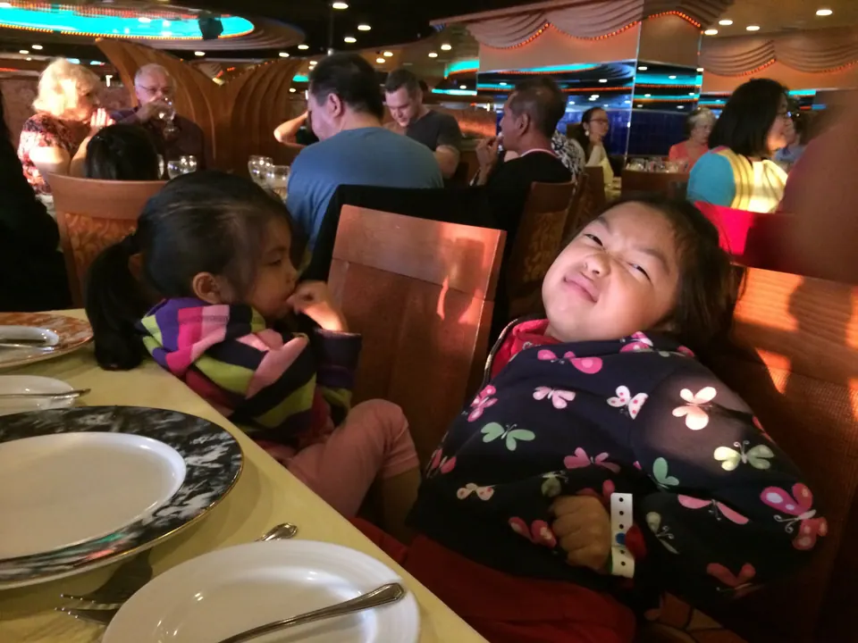 A girl in a butterfly-print jacket tips her head back with her eyes closed, making a comic face, while a smaller girl in stripes eats beside her at a dinner table.