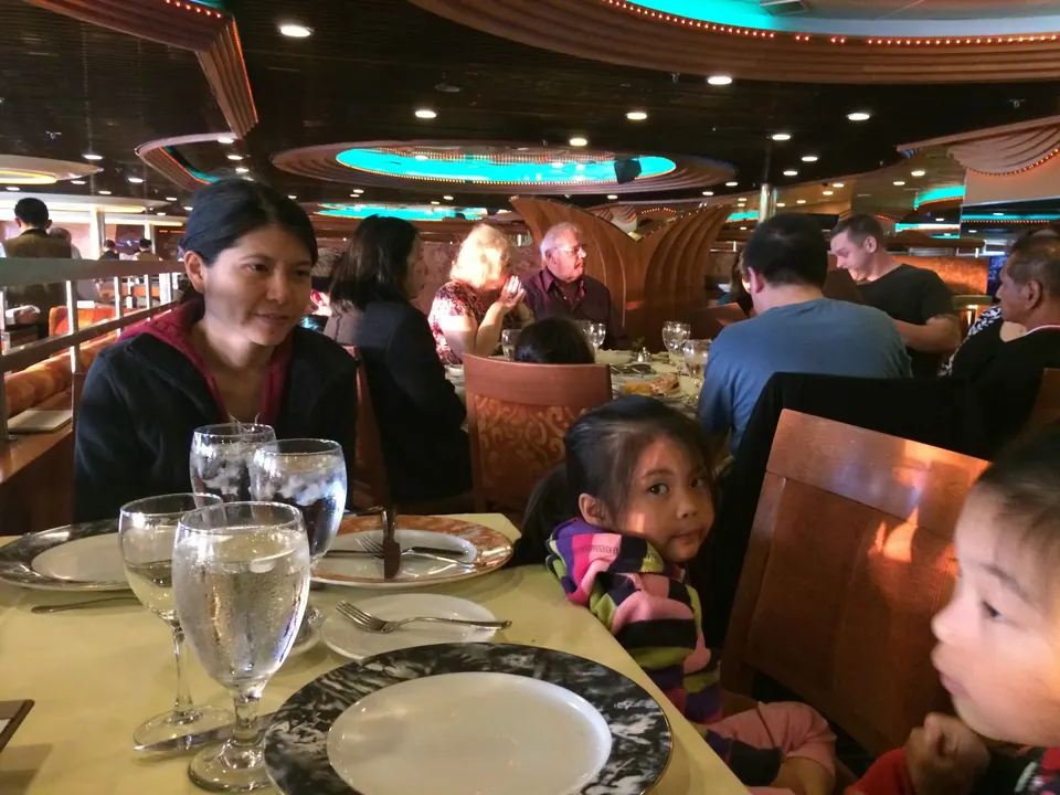 A woman in a black fleece sits behind two water glasses at a dinner table under a turquoise-lit ceiling, with other diners behind her and two girls at the right.