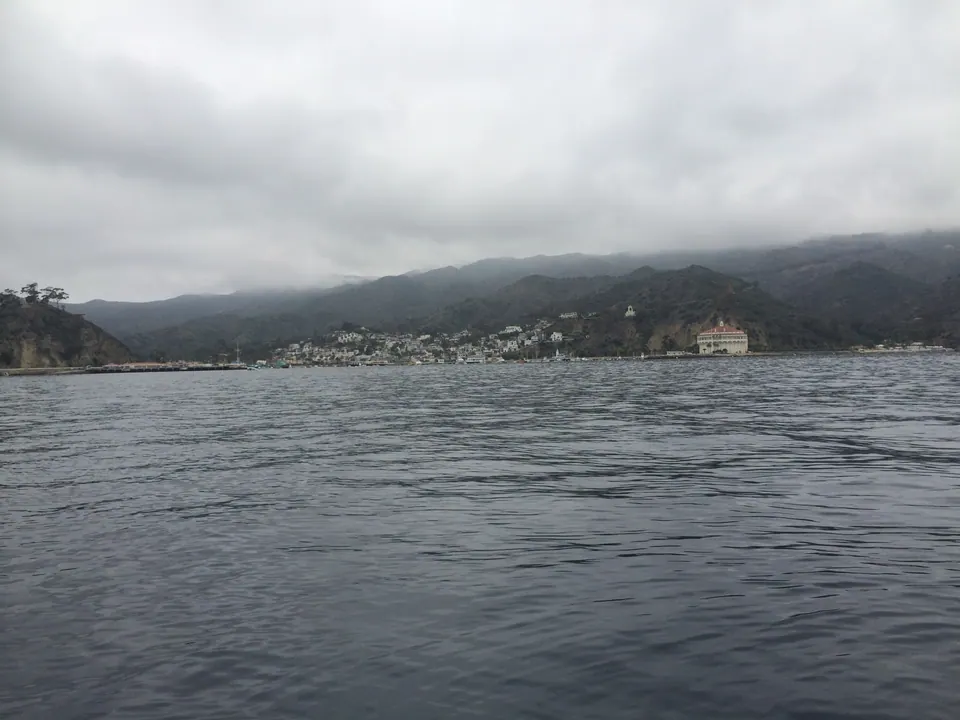 Gray water in front of a small town at the foot of cloud-covered hills; a round white building with a red roof stands on the shore at the right.