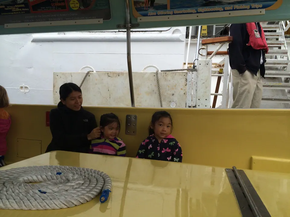 A woman in a black fleece sits close to two small girls on the yellow bench of a tender boat, a coil of white rope in front of them and a crew member in a life vest behind.