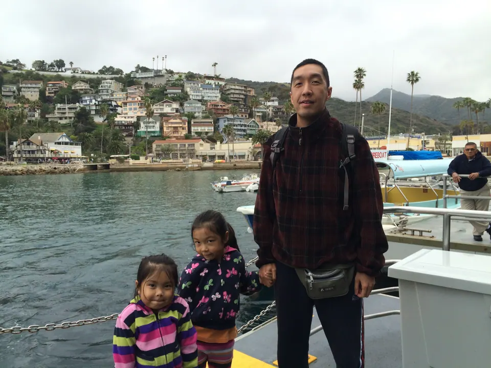 A man in a plaid jacket and a small daypack stands on the tender deck with two small girls; the houses of a hillside town are across the water.