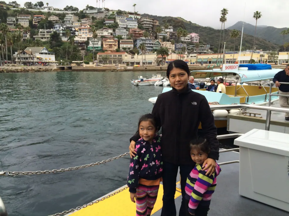 A woman in a black fleece stands with two small girls on the yellow deck of a boat, with moored boats and a hillside of houses and palms behind.