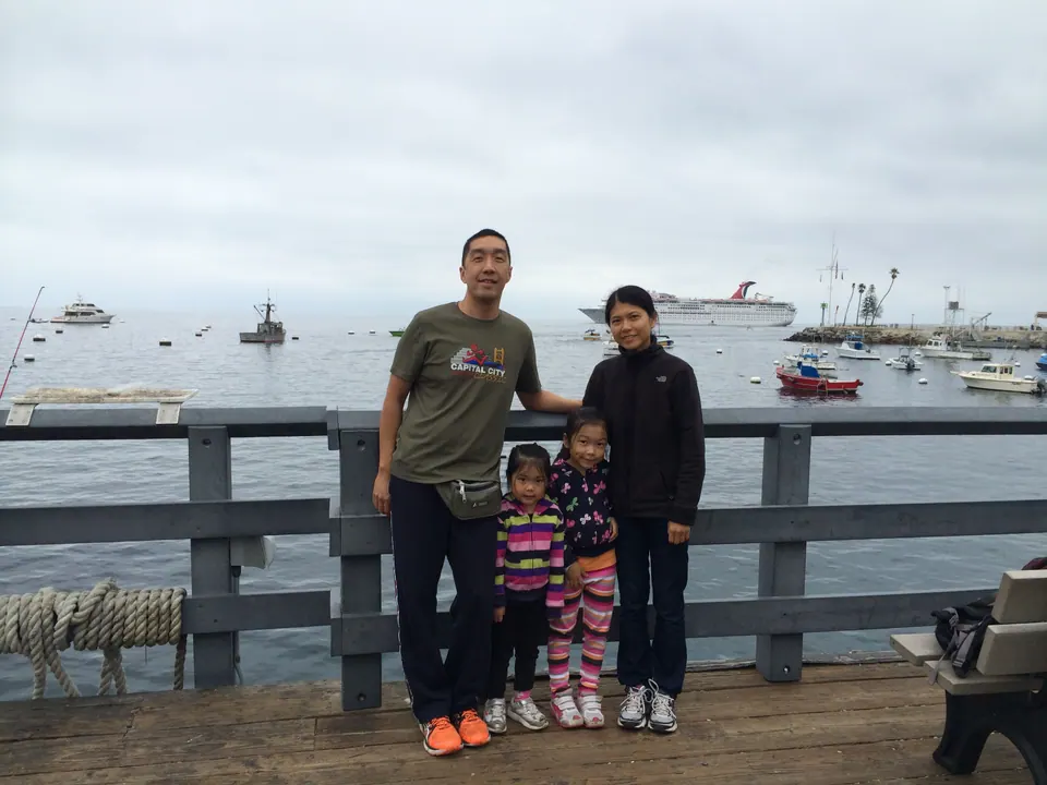 A woman in black, a man in a green T-shirt and two small girls stand in a row at a gray wooden pier railing, the bay and the ship behind them.