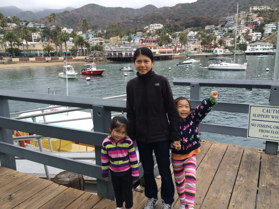 A woman in black stands between a small girl in stripes and a girl in a butterfly-print jacket who raises one arm, on a wooden pier with a No Fishing sign and a colorful hillside town behind.
