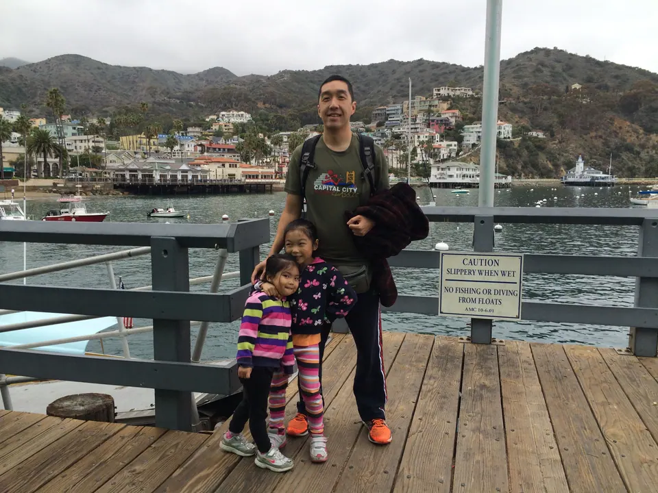 A man with a backpack and two small girls stand on a wooden pier by a railing, with the harbor, boats and a hillside town behind them.