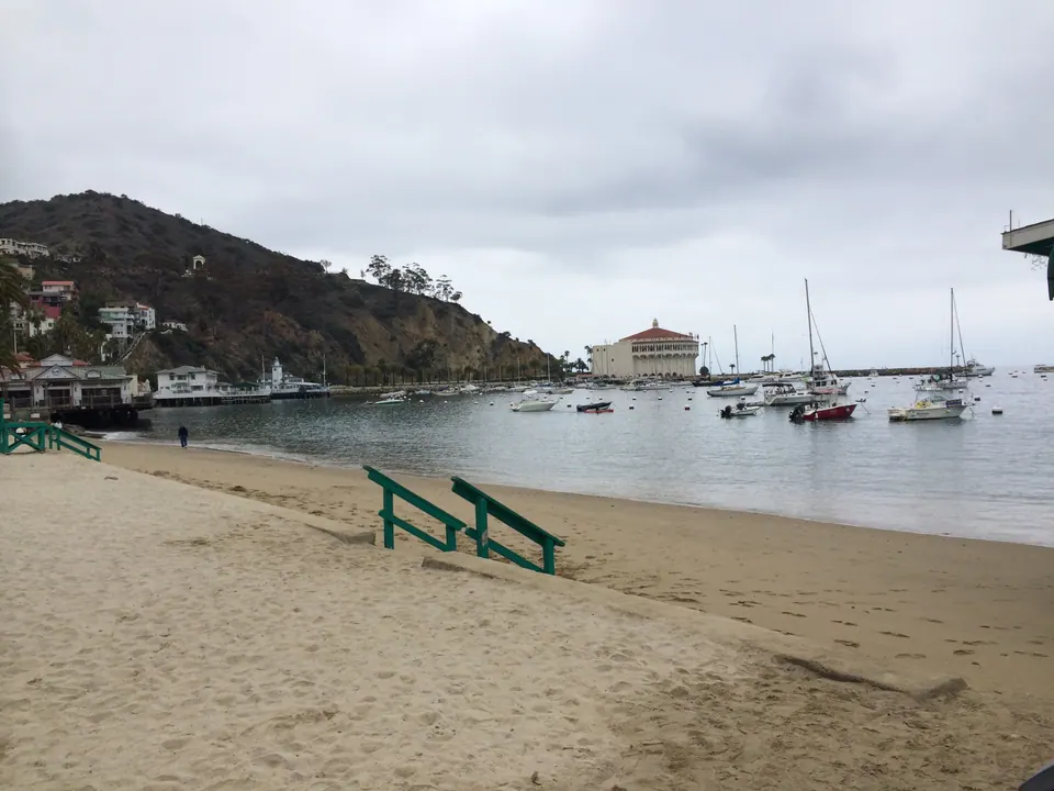 An empty sand beach with green railings curves toward a bay full of moored sailboats, with a round building on the far point under a gray sky.