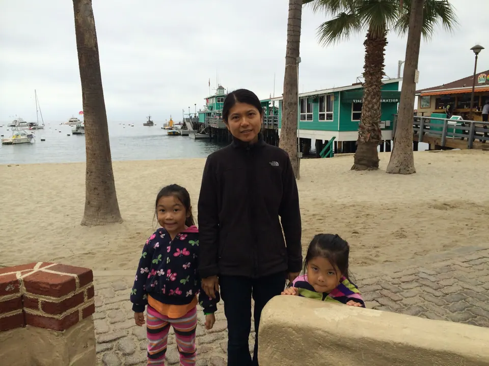 A woman in black stands between two small girls on a paved promenade beside a brick planter, with palms and teal pier buildings behind them.