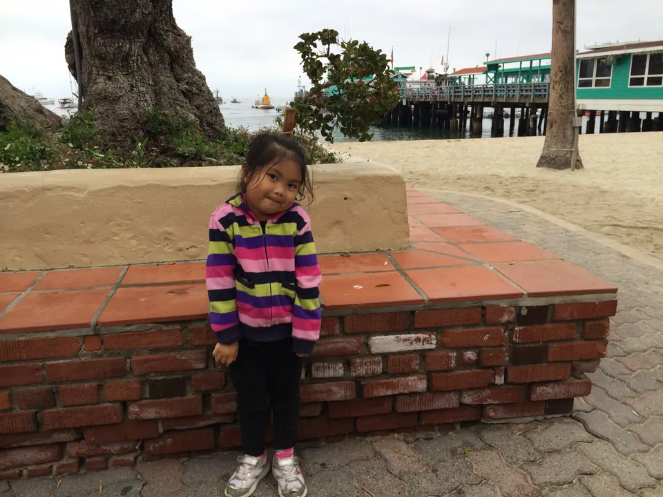 A small girl in a striped jacket stands with her head tipped in front of a brick planter, the pier and moored boats behind her.