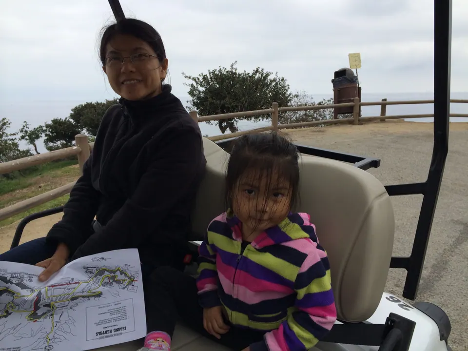 A woman holding a paper map sits in a golf cart beside a small girl in a striped jacket, with a fence and the sea behind them.