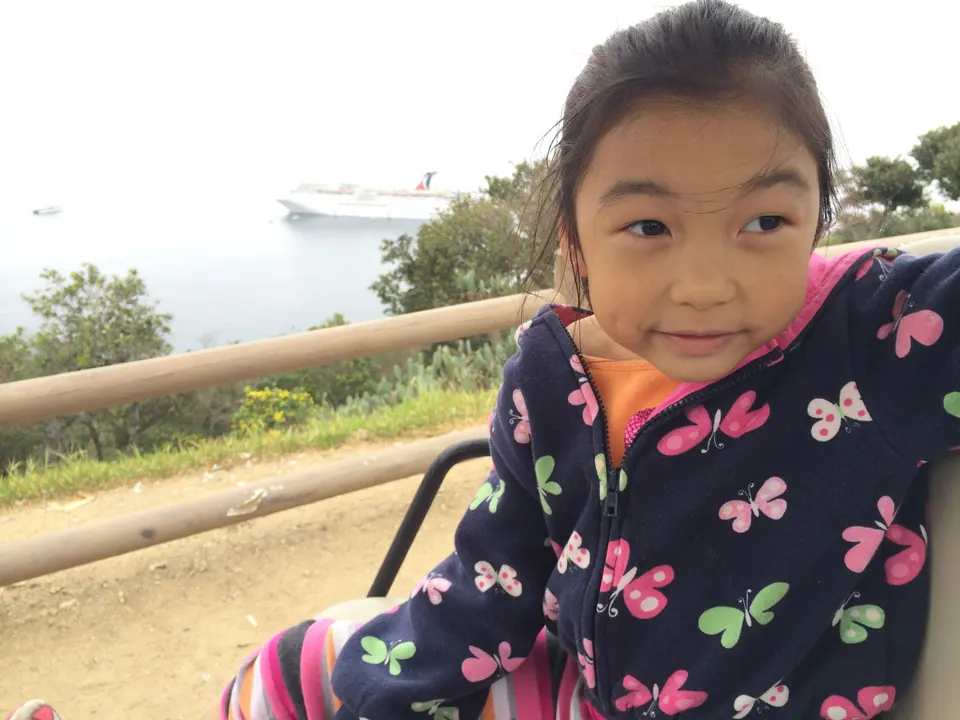 A close-up of a girl in a butterfly-print jacket in a golf cart, with a wooden rail and a small white ship on the water behind her.