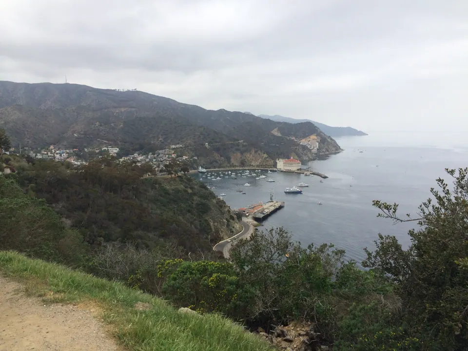 From a hillside above a bay, a harbor with a pier, moored boats and a round Casino building on the point below dark, cloud-topped ridges.