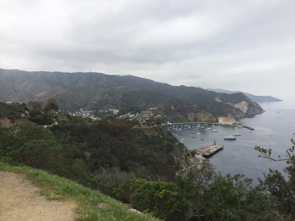 A wide view from a hill road over a bay, town and the Casino point, the mountainous coast running out to sea under a gray sky.