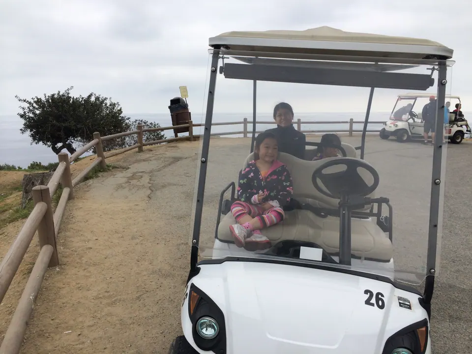 A white golf cart numbered 26 stopped at a hill overlook with a girl in the front seat and riders behind her, beside a wooden fence at the cliff edge.
