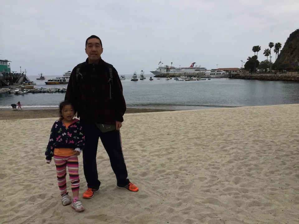 A man and a small girl stand on a sandy beach; boats and a large white cruise ship are on the water behind them.