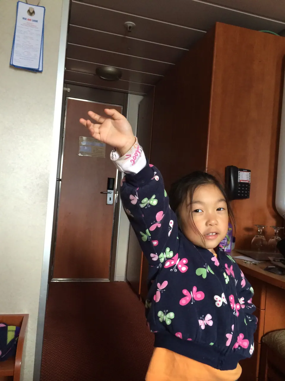 A girl in a butterfly-print jacket stands in a cabin with one arm raised high, a door, a phone and wood cabinets behind her.