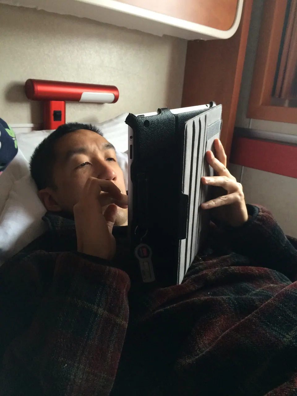 A man in a plaid shirt lies on a bed holding a tablet up over his face, with a red reading lamp above him.