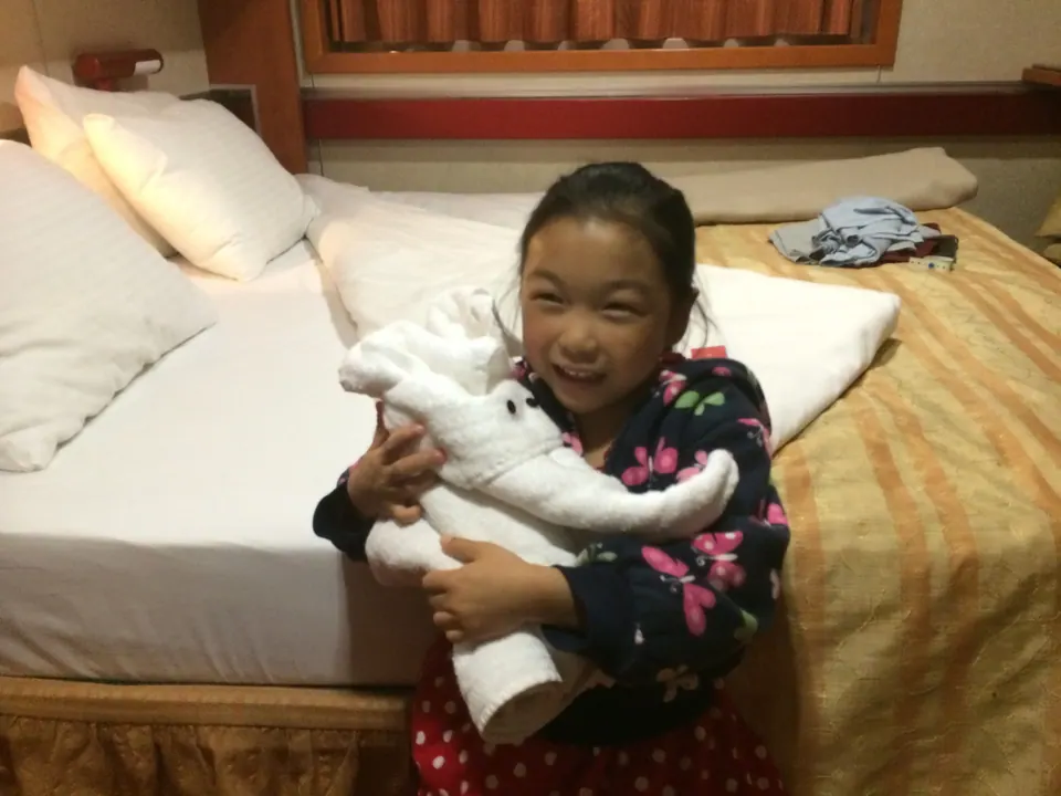 A laughing girl in a butterfly-print jacket hugs a towel elephant on the edge of a bed with a striped bedspread.