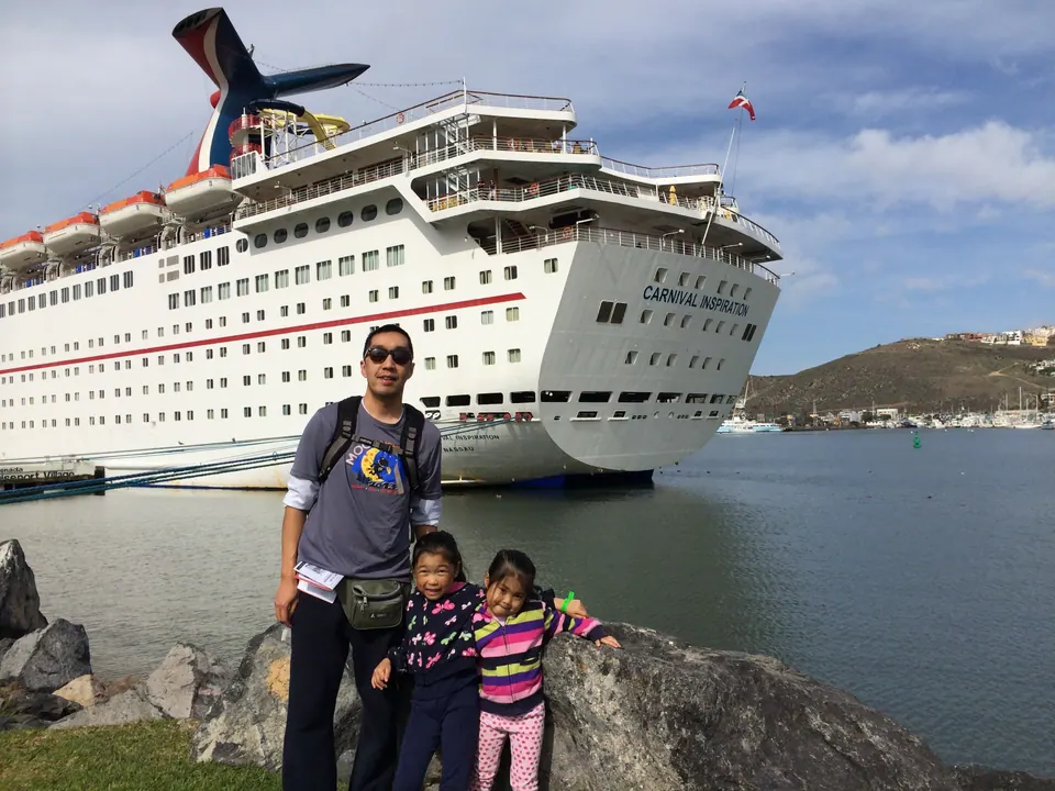 A man in sunglasses and two small girls stand on rocks at the water's edge in front of the bow of a large white cruise ship named Carnival Inspiration, under a blue sky with broken cloud.