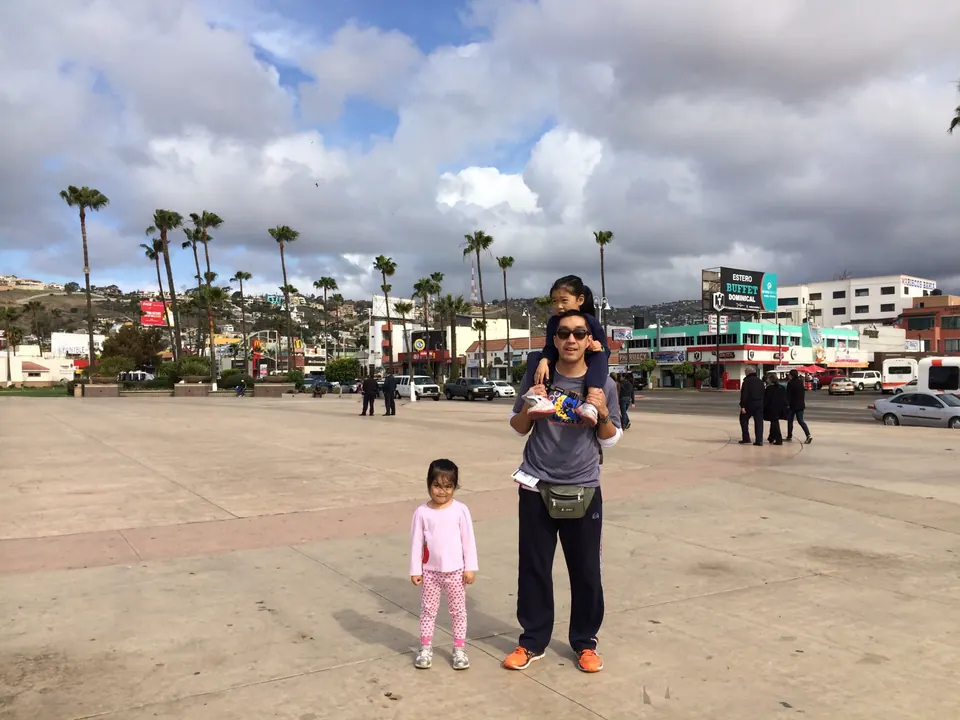 A man carries a girl on his shoulders on a wide paved plaza lined with tall palms and shops, while a smaller girl in pink stands nearby, under big white clouds.