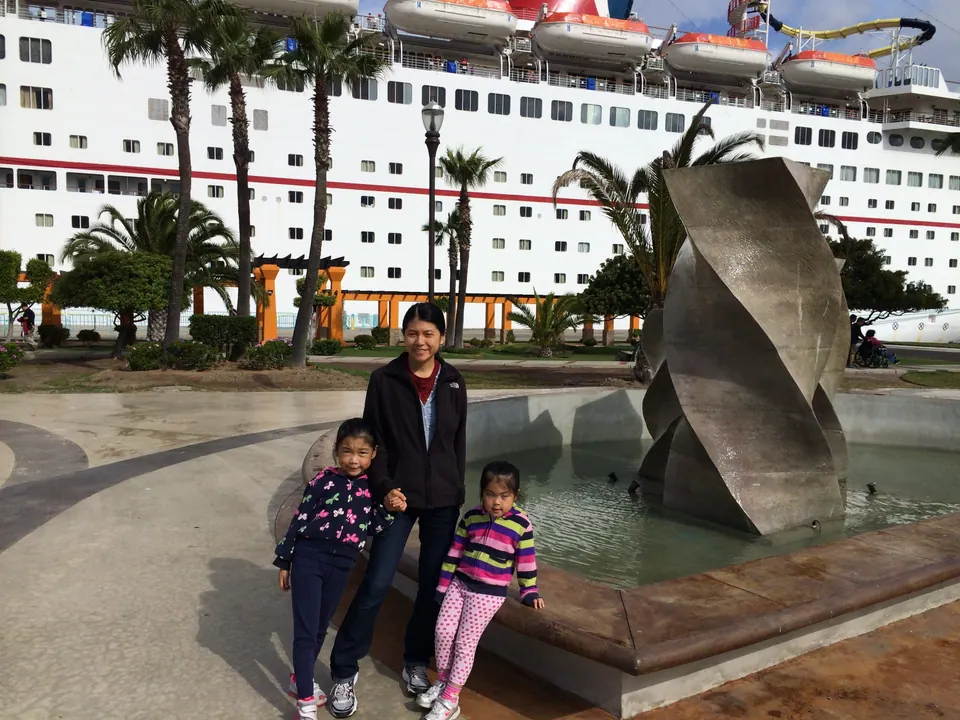 A woman in a black fleece and two small girls stand at the edge of a fountain with a twisted bronze sculpture, with the white side of a cruise ship and palm trees rising behind them.