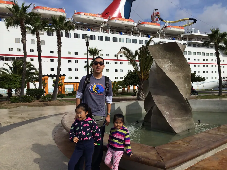 A man in sunglasses and a daypack stands with two small girls at a fountain with a twisted sculpture, palms and the ship's lifeboats and funnel above them.