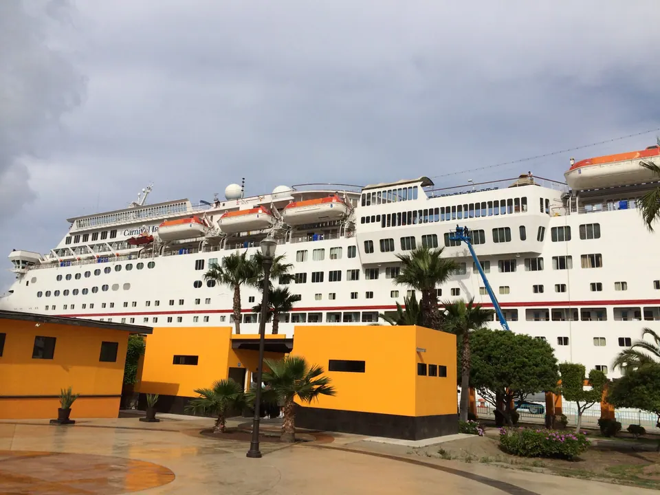 A large white cruise ship rises behind palm trees and a low orange building under a blue sky with broken cloud.