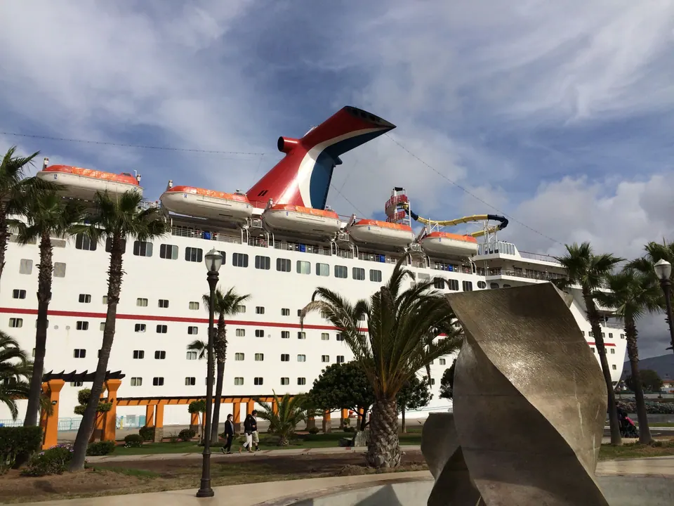 Looking up past a curved bronze sculpture and palm trees at the white side of a cruise ship and its red, white and blue winged funnel against a bright sky.