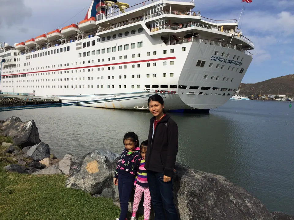 A woman in a black fleece and two small girls stand on rocks by the water in front of the stern of the Carnival Inspiration, with hills across the harbor.