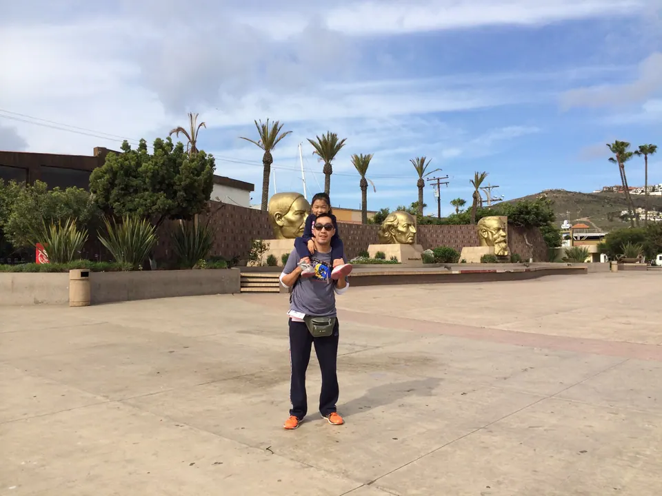 A man with a girl on his shoulders stands in the middle of a huge empty paved plaza; a row of palms and large golden faces on a wall are behind them.