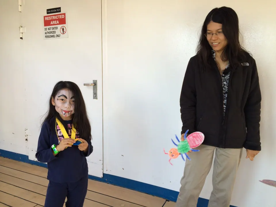 A girl with black and white face paint and a yellow lanyard stands by a white door on a wooden deck; a woman beside her holds a colored paper bug.